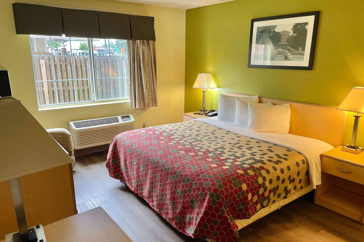 Une chambre d’hôtel avec un lit, une table de chevet, une lampe, une fenêtre avec des rideaux et un tableau encadré au mur.