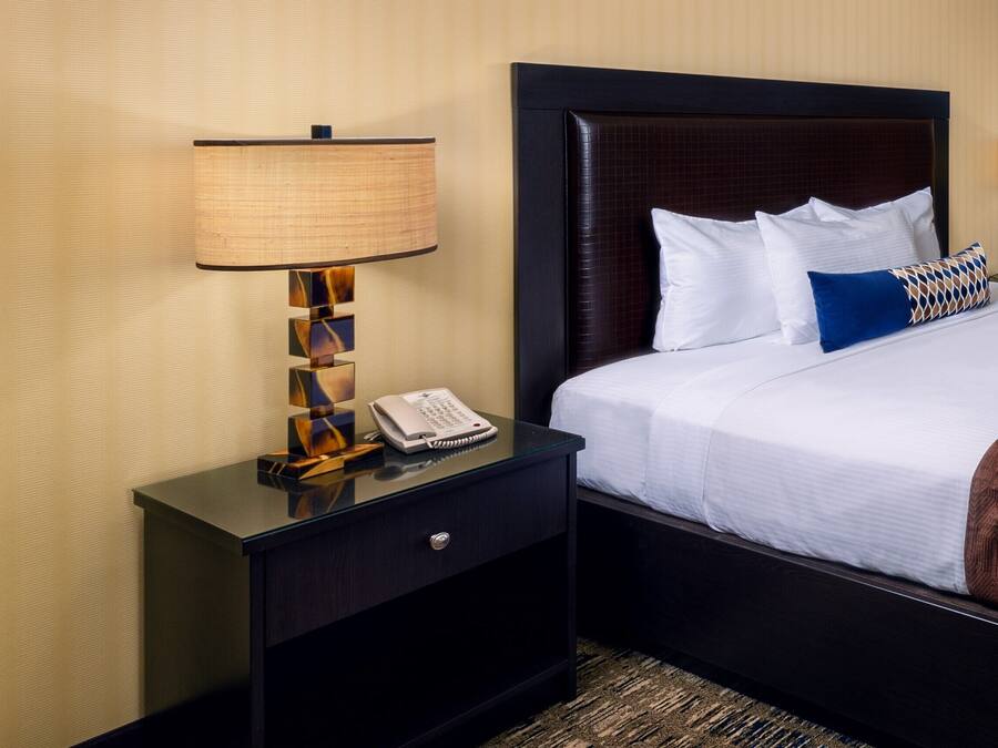 A hotel room with a bed, a nightstand with a lamp, a telephone, and a brown throw blanket.