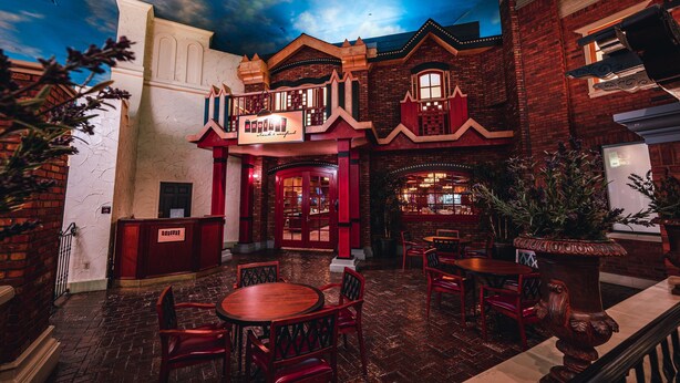 Un edificio de ladrillo con entrada roja, balcón y zona de bar con mesas y sillas.