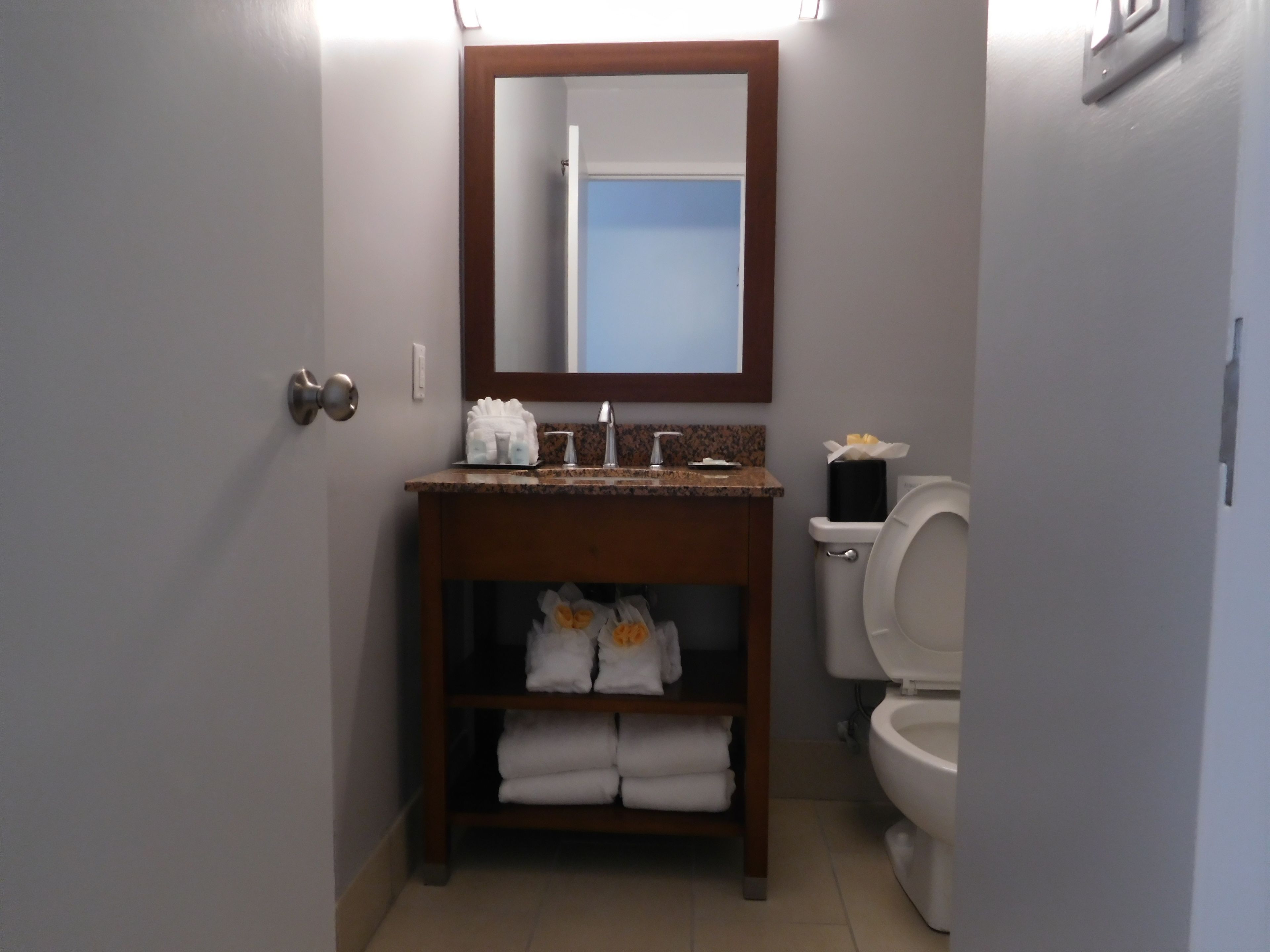 A bathroom with a wooden vanity, a mirror, a toilet, and a shelf with folded towels.