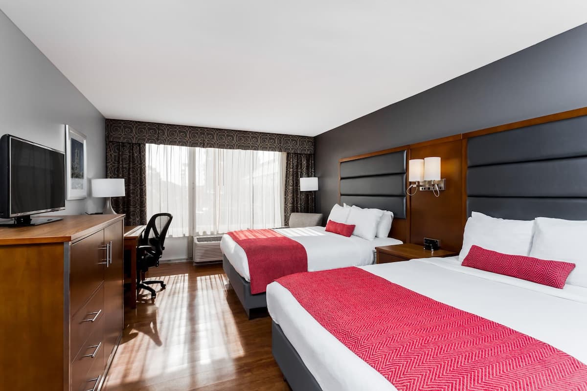 Ein Hotelzimmer mit zwei Betten, einem Schreibtisch mit Fernseher, einem Sessel, einer Lampe und einem Fenster mit Vorhängen.