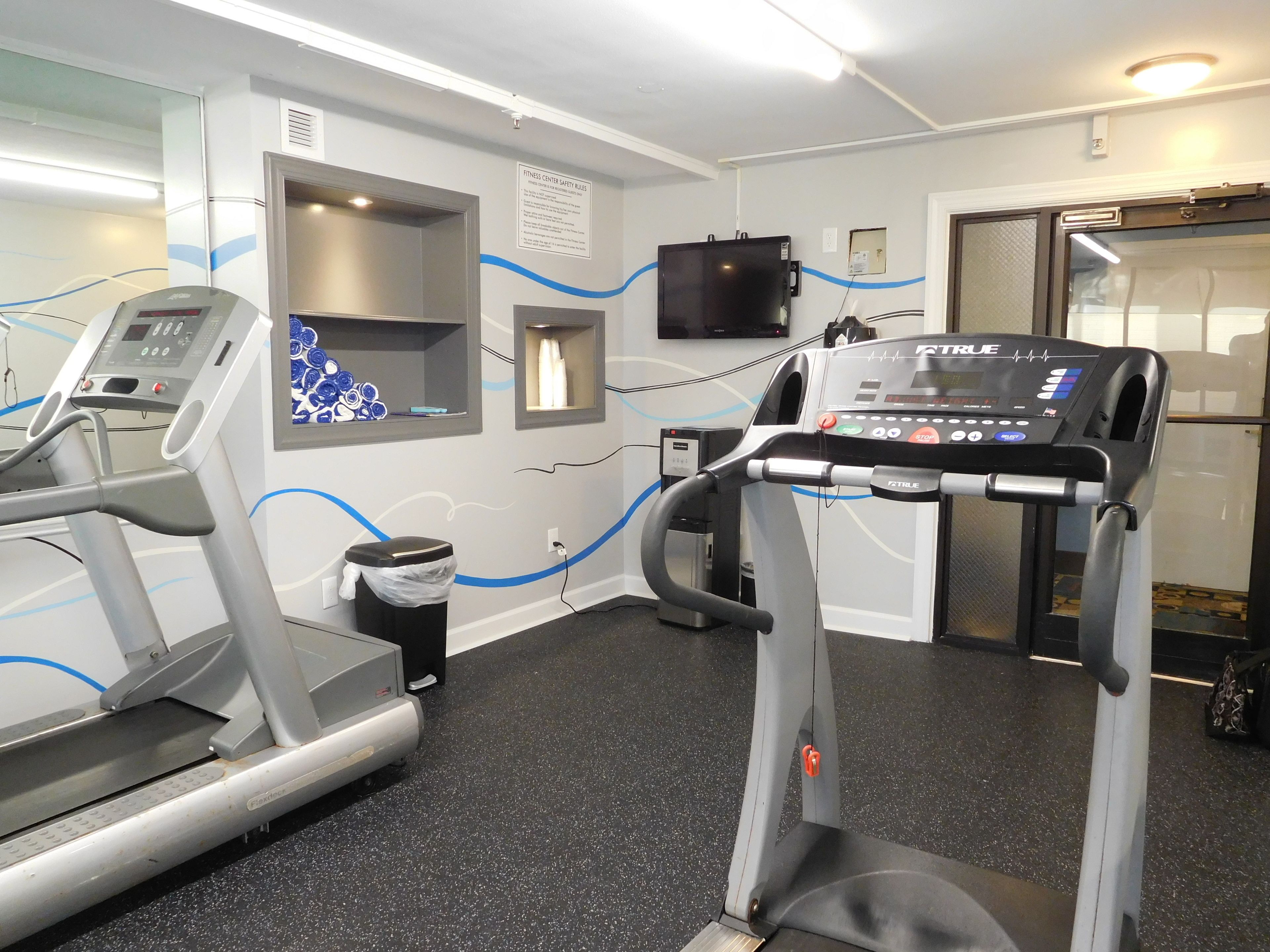 Un gimnasio con cintas de correr, un televisor montado en la pared y un cubo de basura.