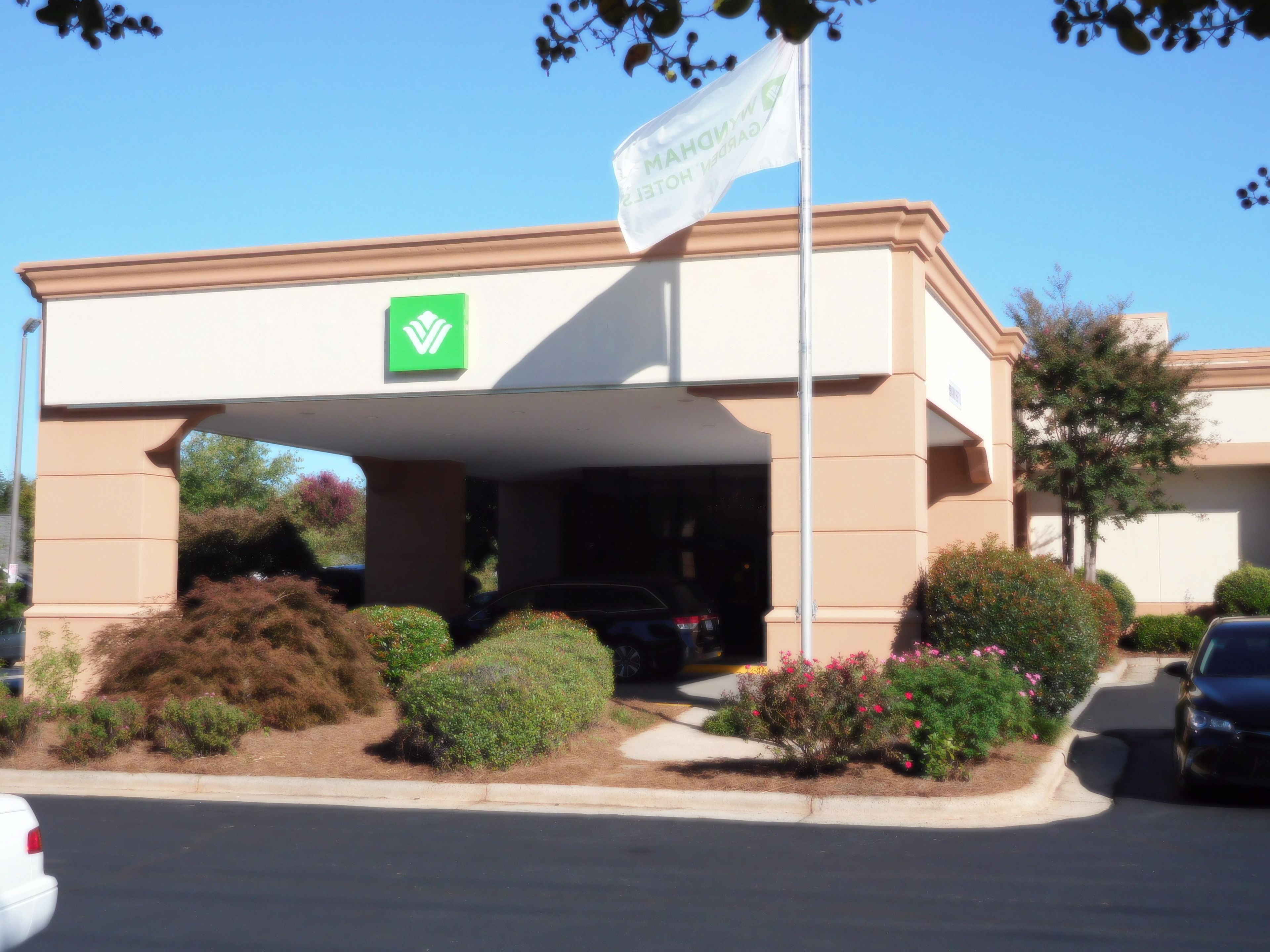Entrada de un hotel con un logotipo verde, una bandera y un estacionamiento con coches y jardines.