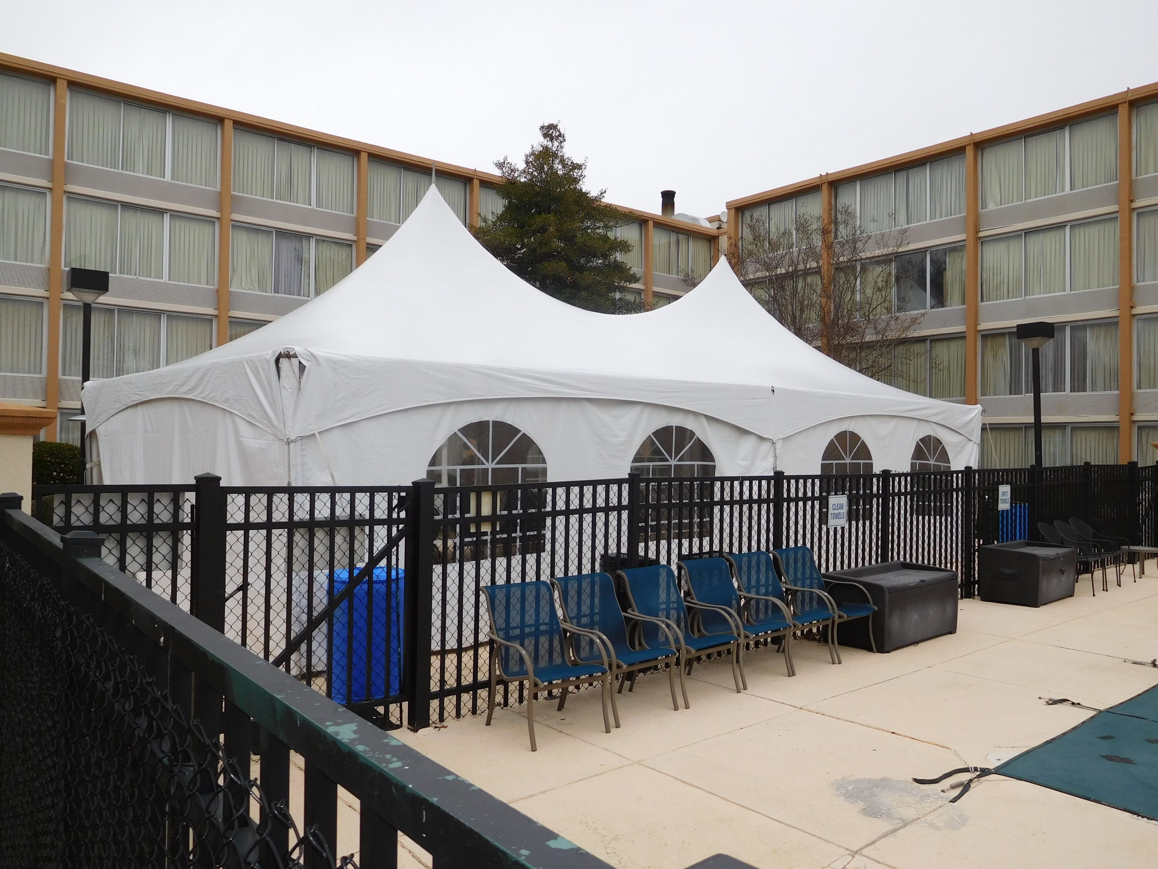 A white tent with arched windows, blue chairs, and a black fence in front of a building with large windows and a brick facade.