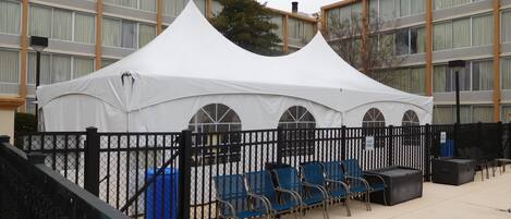 A white tent with arched windows, blue chairs, and a black fence in front of a building with large windows and a brick facade.