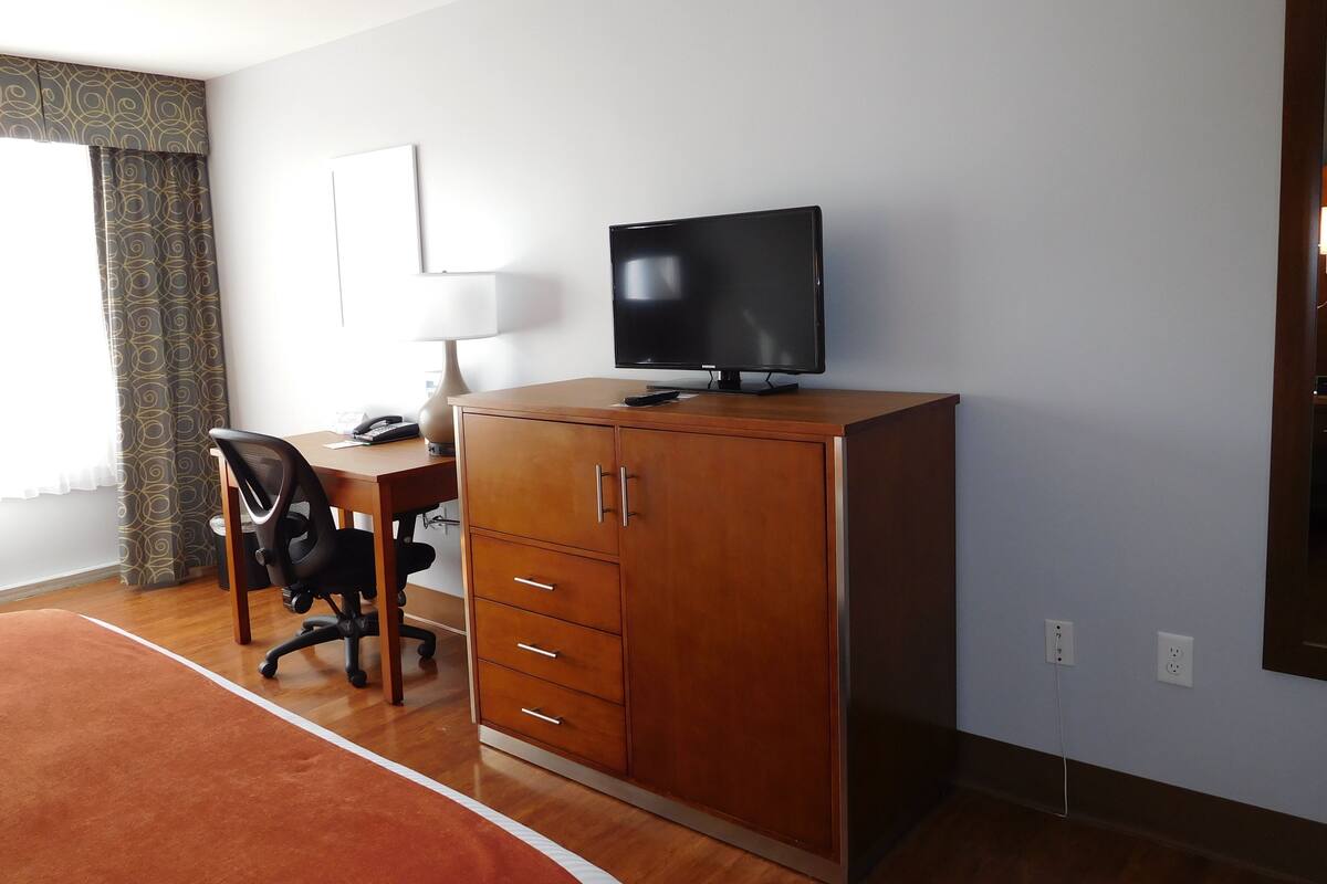 Ein Hotelzimmer mit einem Holztisch, einem schwarzen Bürostuhl, einem Fernseher auf einem Schrank, einer Lampe und einem fenster mit Vorhängen.