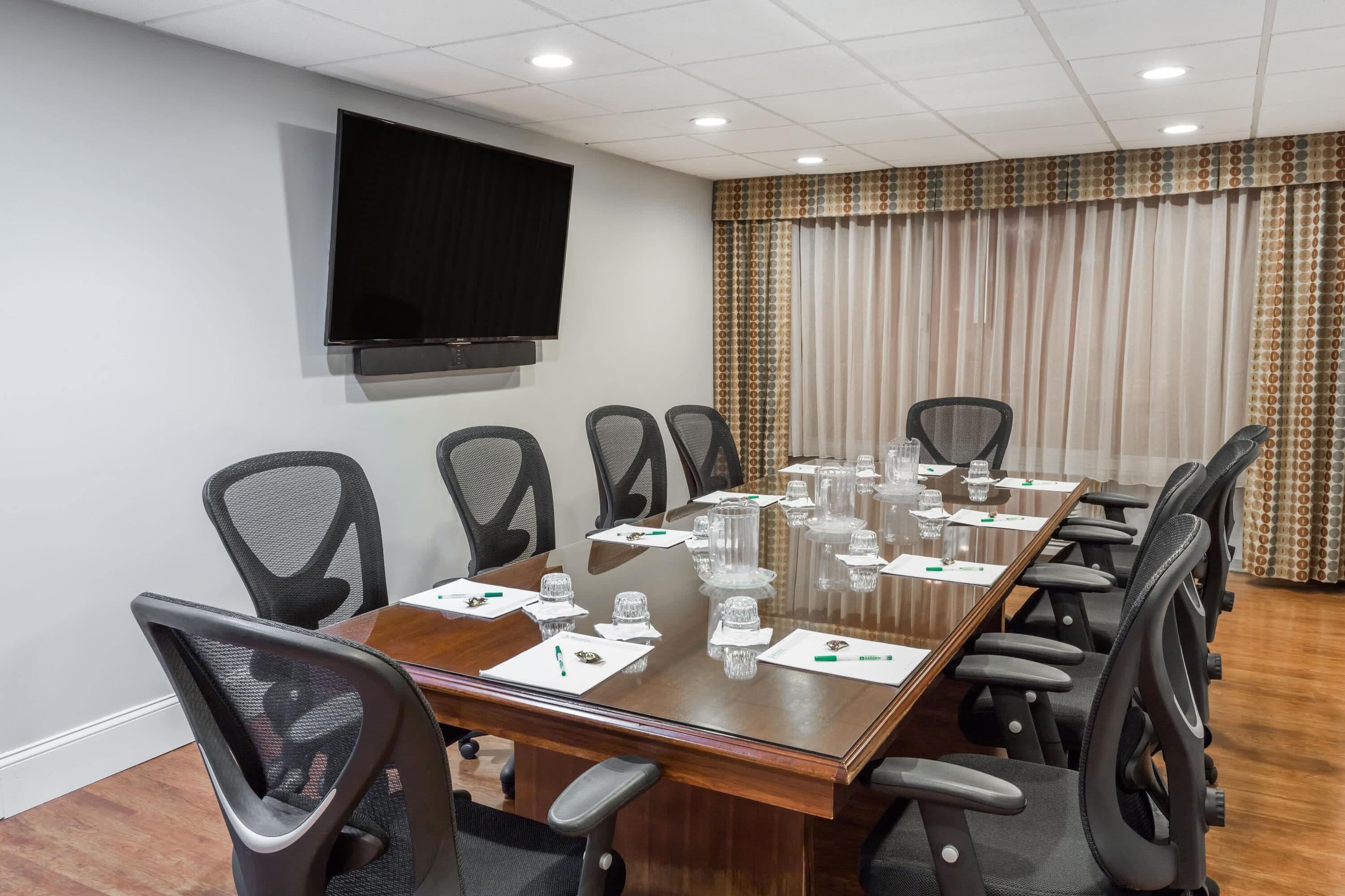 A conference room with a long table, multiple chairs, a flat-screen TV, and a patterned curtain.