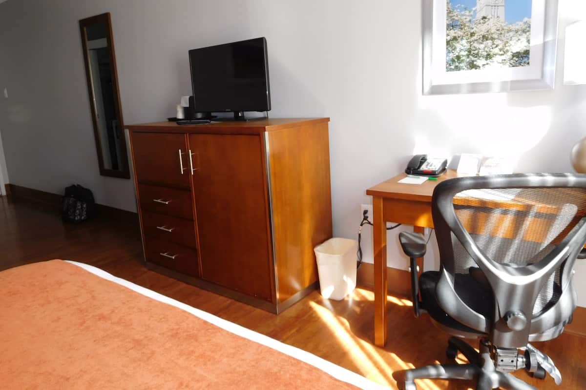 Ein Zimmer mit einem Holztisch, einem schwarzen Bürostuhl, einem Fernseher auf einem Schrank, einem Fenster mit Blick auf ein Gebäude und einem Mülleimer.