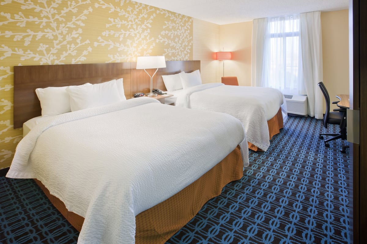 Une chambre d’hôtel avec deux lits, un bureau, une chaise, une lampe et une fenêtre avec des rideaux.