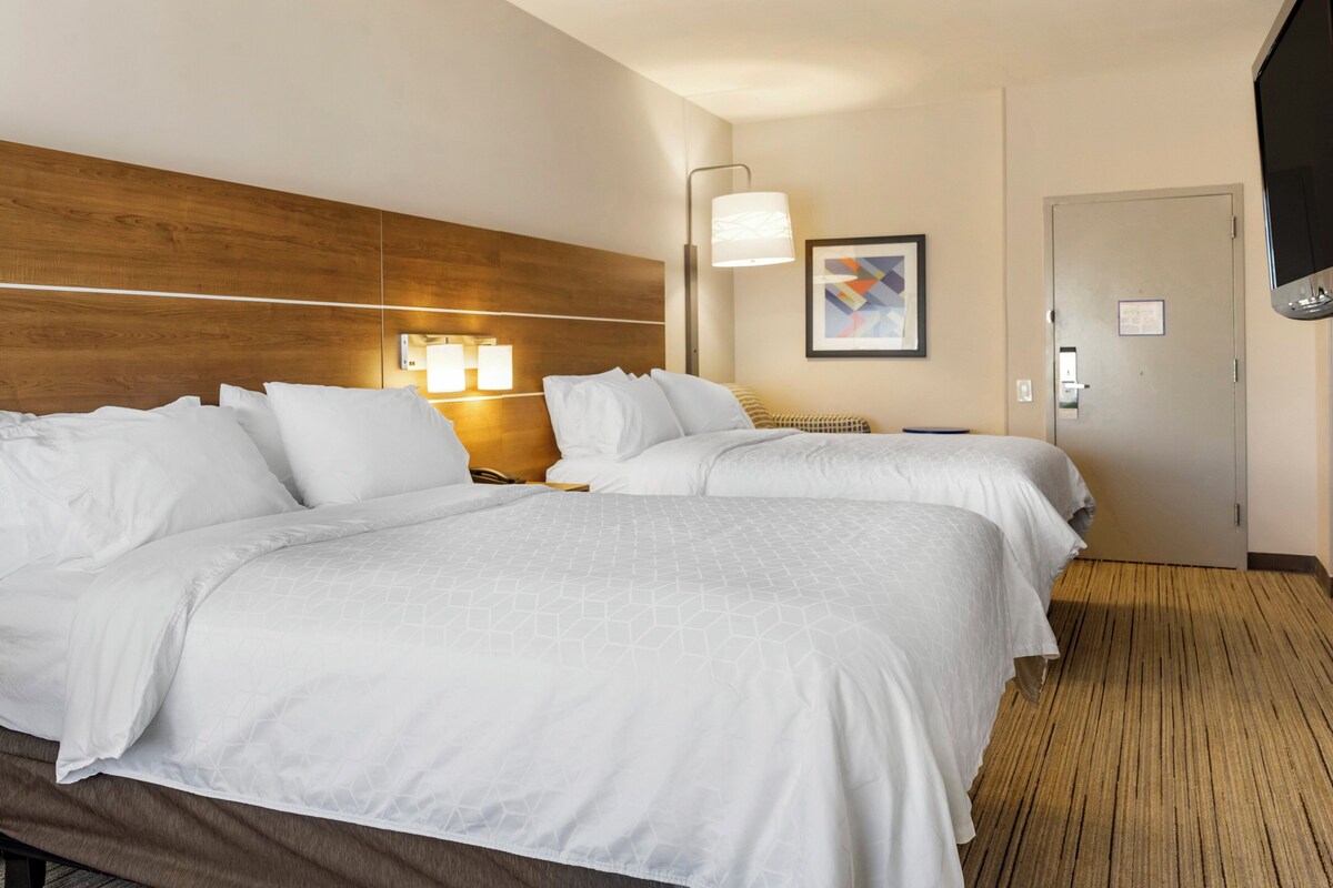 A hotel room with two beds, a wooden headboard, a wall-mounted TV, a framed abstract painting, and a door with a sign.