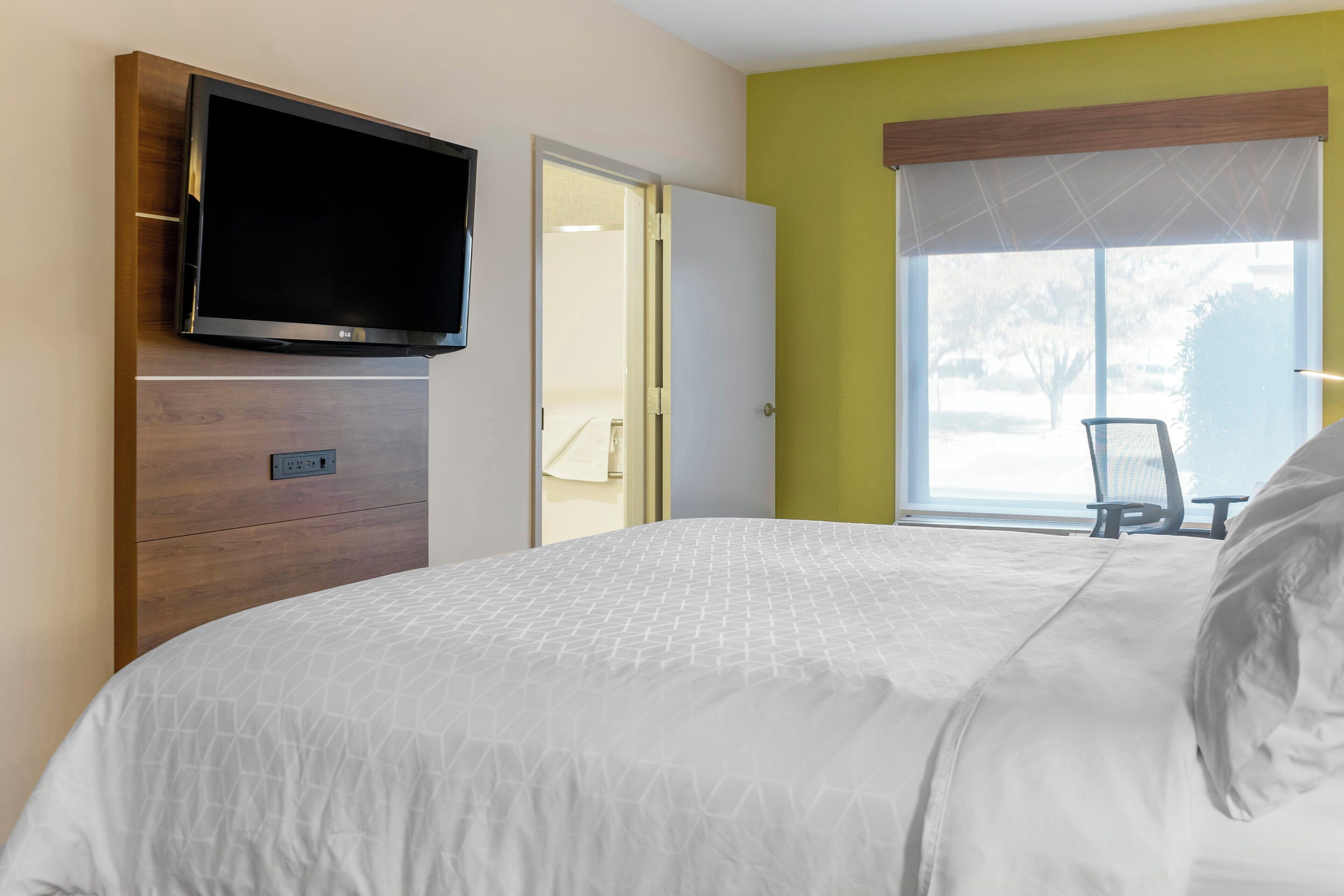 Una habitación de hotel con una cama, un televisor montado en una cómoda de madera, una ventana con vistas y una puerta que da a otra habitación.
