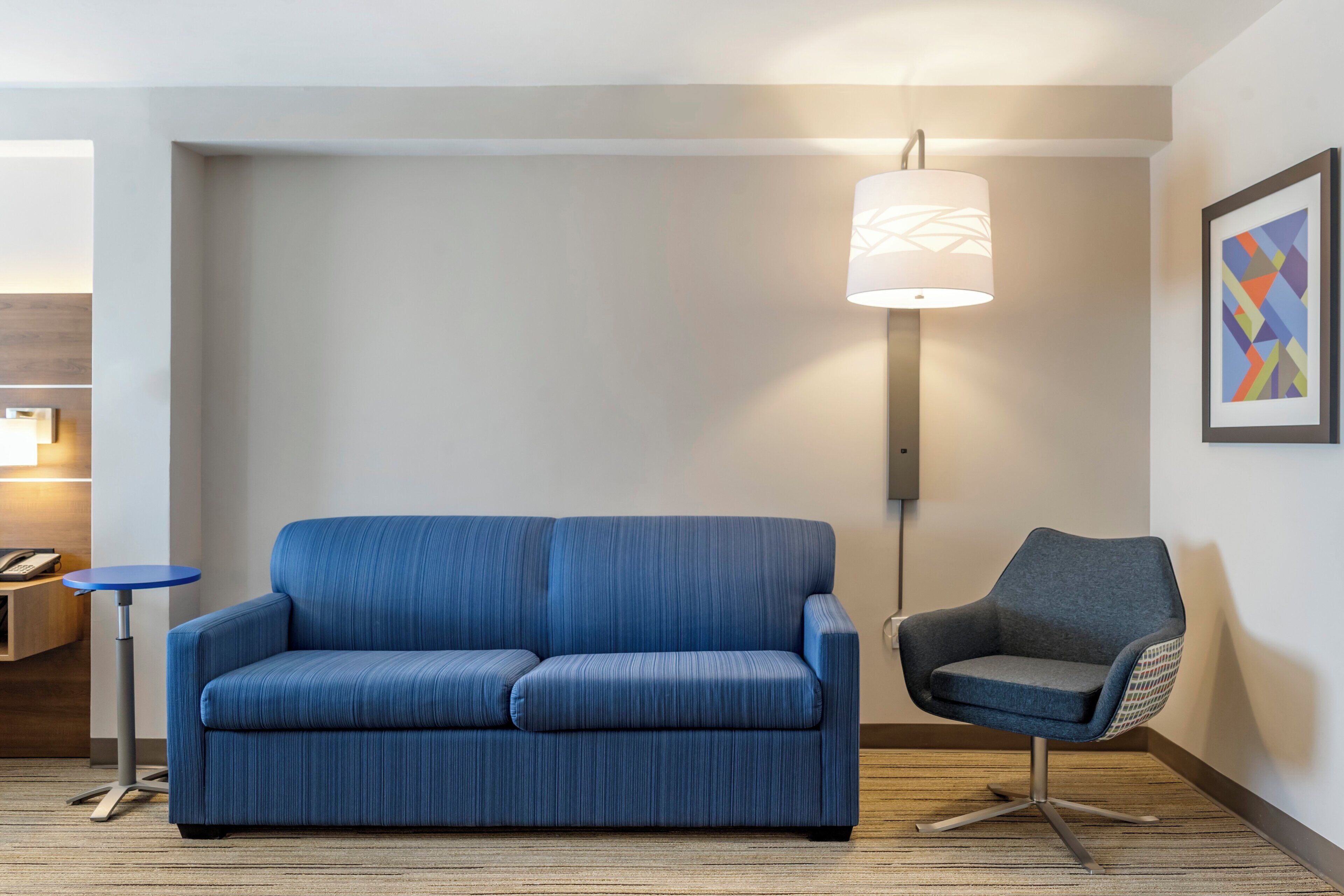 Een moderne woonkamer met een blauwe bank, een grijze fauteuil, een vloerlamp en een lijst met een abstract schilderij aan de muur.