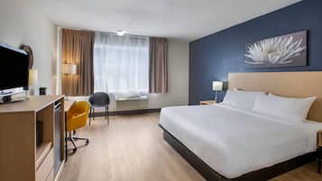A hotel room with a large bed, a desk with a computer, a chair, a window with curtains, and a wall-mounted lamp.