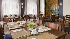 Un comedor de hotel con varias mesas dispuestas para el desayuno, con posavasos, tazas y un centro de mesa con planta decorativa.