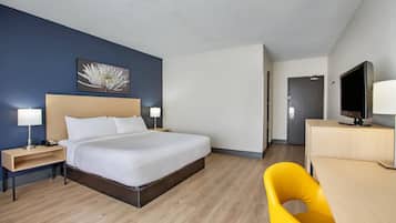 Una habitación de hotel con cama, mesitas de noche, un escritorio con silla, televisor y puerta.