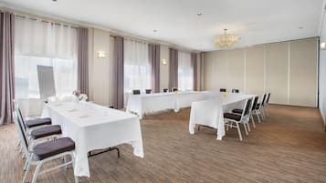 A conference room with multiple tables and chairs, large windows with curtains, and a chandelier.