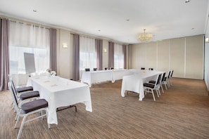 Une salle de conférence dotée de plusieurs tables et de chaises, de grandes fenêtres avec des rideaux et d’un lustre.