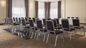 Un salón de conferencias con filas de sillas, ventanas amplias con cortinas y una araña.