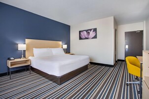 A hotel room with a large bed, a nightstand with a lamp, a desk with a chair, and a door.