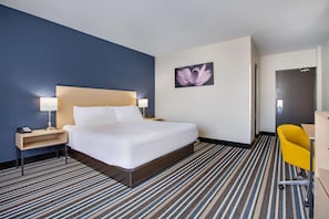 Une chambre d’hôtel avec un grand lit, une table de chevet avec une lampe, un bureau avec une chaise et une porte.