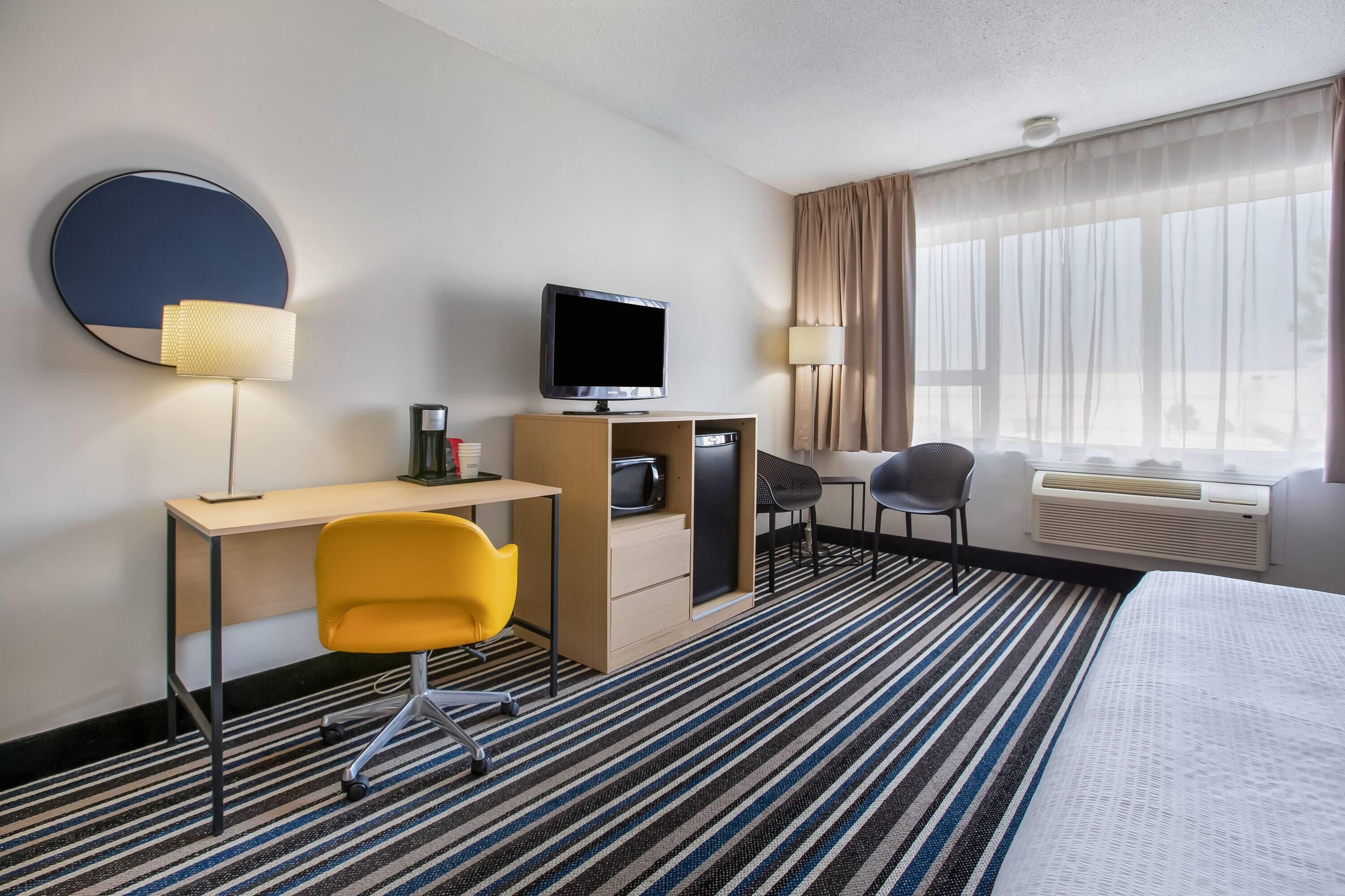 A hotel room with a bed, a desk with a chair, a television, a coffee machine, and a window with curtains.