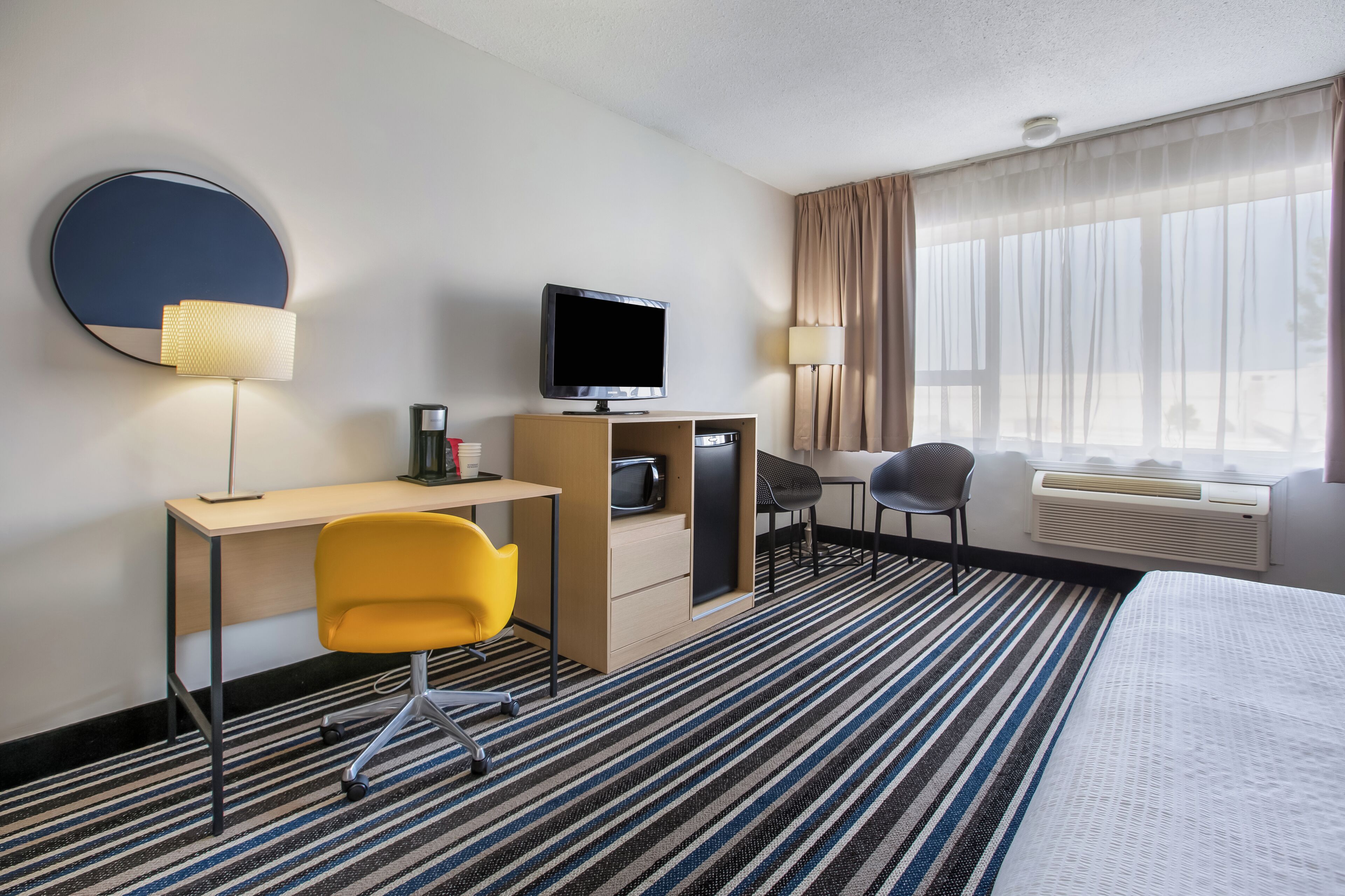 Habitación de hotel con cama, escritorio, silla, televisor, cafetera y ventana con cortinas.