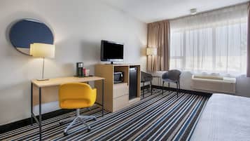 Habitación de hotel con cama, escritorio, silla, televisor, cafetera y ventana con cortinas.