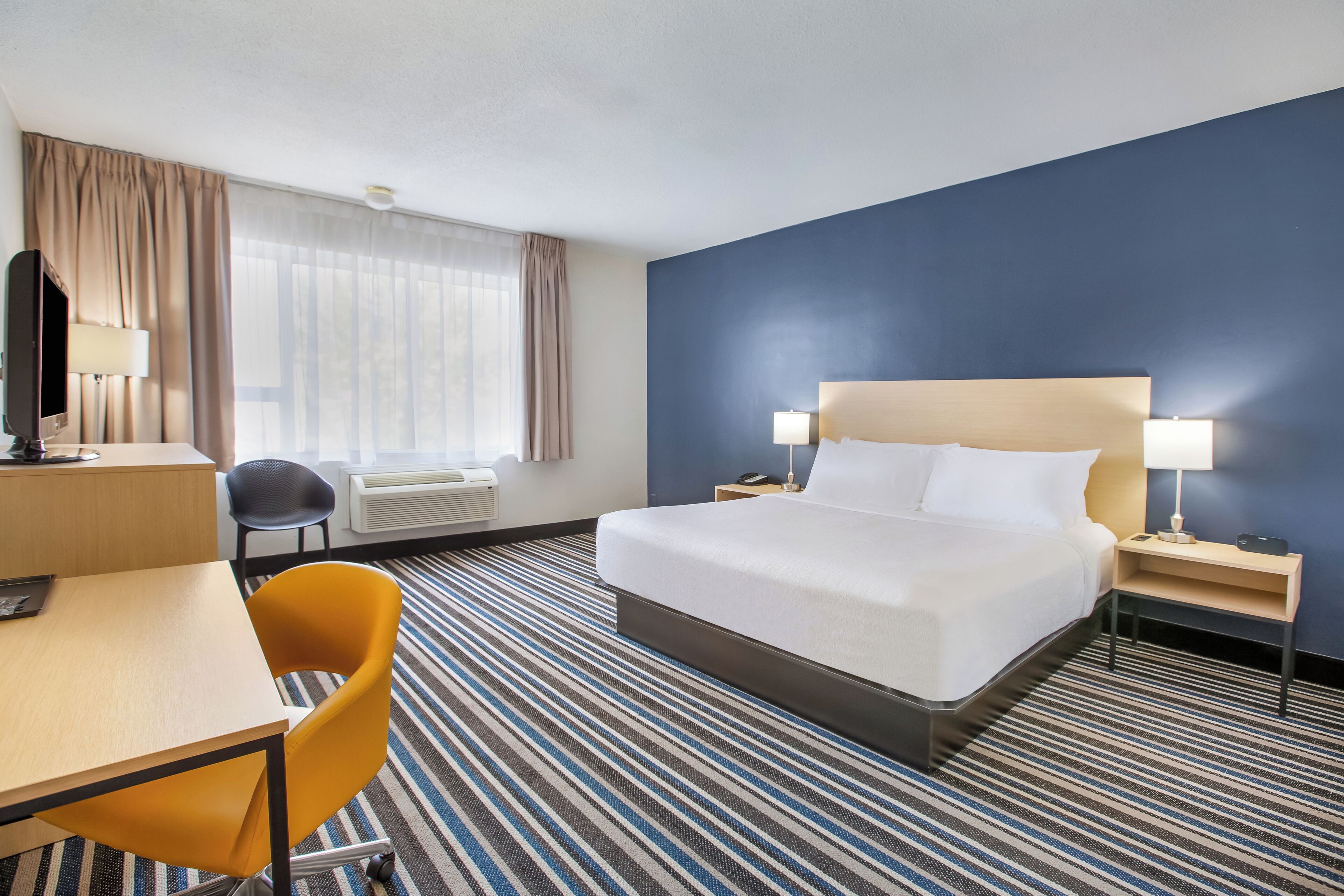 Ein Hotelzimmer mit einem Bett, einem Schreibtisch, einem Stuhl, einer Schreibtischlampe, einem Nachttisch mit Lampe und einem Fenster mit Vorhängen.