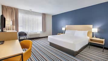 Ein Hotelzimmer mit einem Bett, einem Schreibtisch, einem Stuhl, einer Schreibtischlampe, einem Nachttisch mit Lampe und einem Fenster mit Vorhängen.