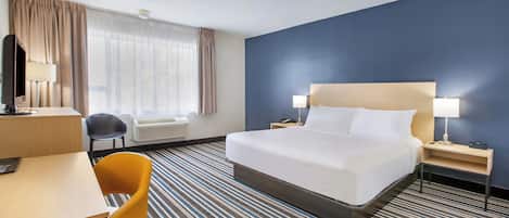 Habitación de hotel con cama, escritorio, silla, lámpara de escritorio, mesita de noche con lámpara y ventana con cortinas.