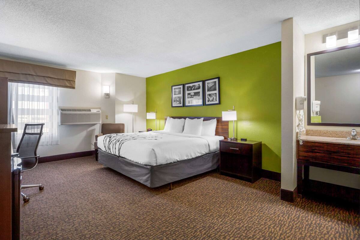 Quarto de hotel com cama, mesas de cabeceira, escrivaninha, cadeira, espelho e ar condicionado fixado na parede.