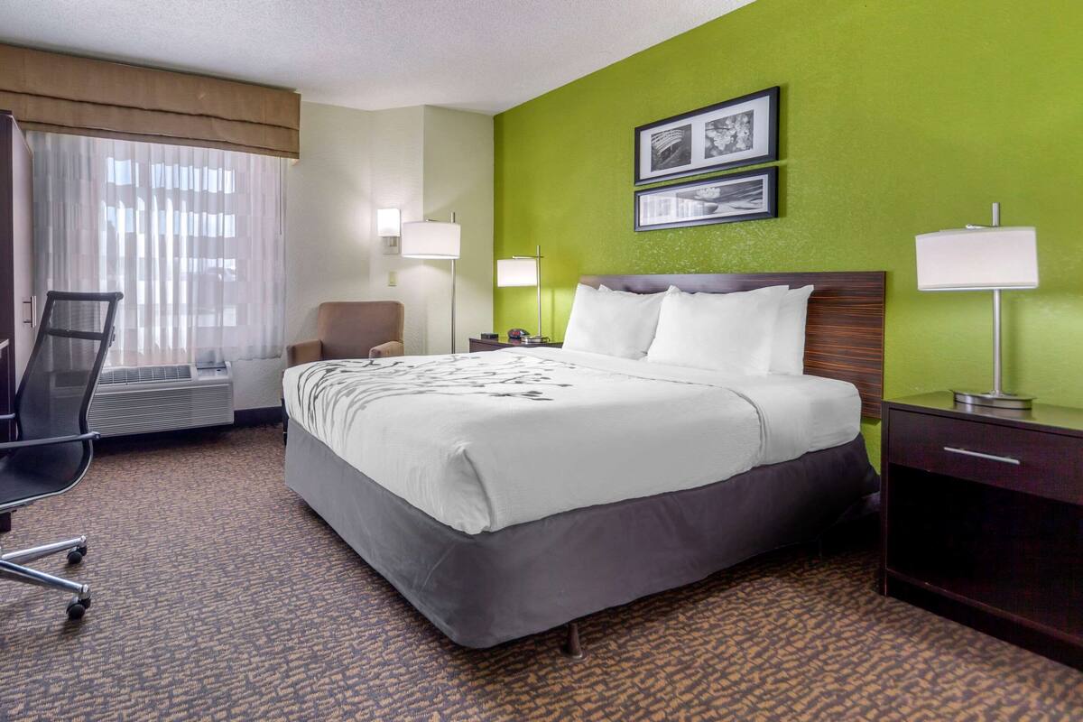 Quarto de hotel com uma cama grande, uma escrivaninha, uma cadeira e uma luminária de mesa.