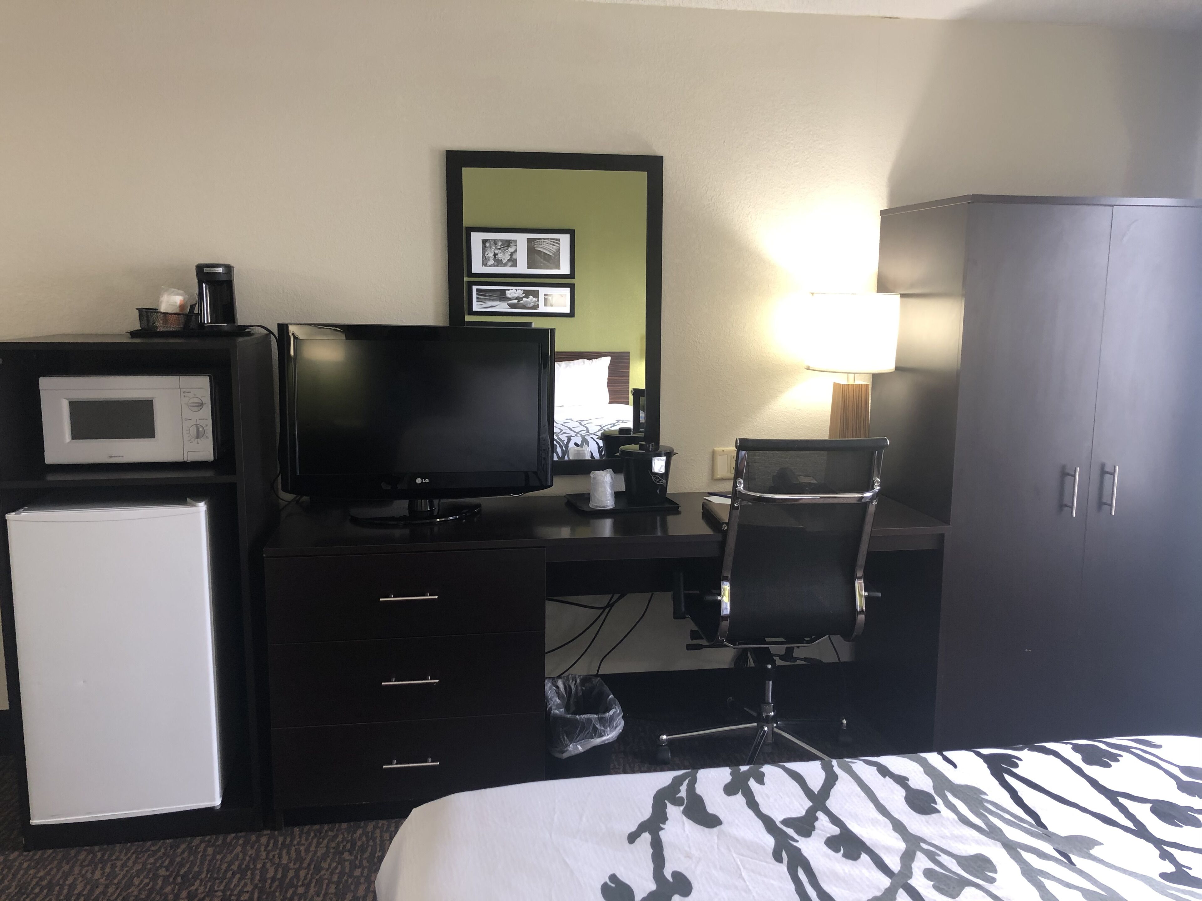 A hotel room with a bed, a desk with a computer, a microwave, a refrigerator, a chair, and a wardrobe.