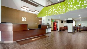 A hotel reception area with a wooden reception desk, a green wall with a leaf pattern, and a seating area with tables and chairs.