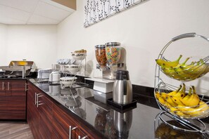 A modern kitchen with a stainless steel coffee station, a fruit basket, and a buffet area with various food items.
