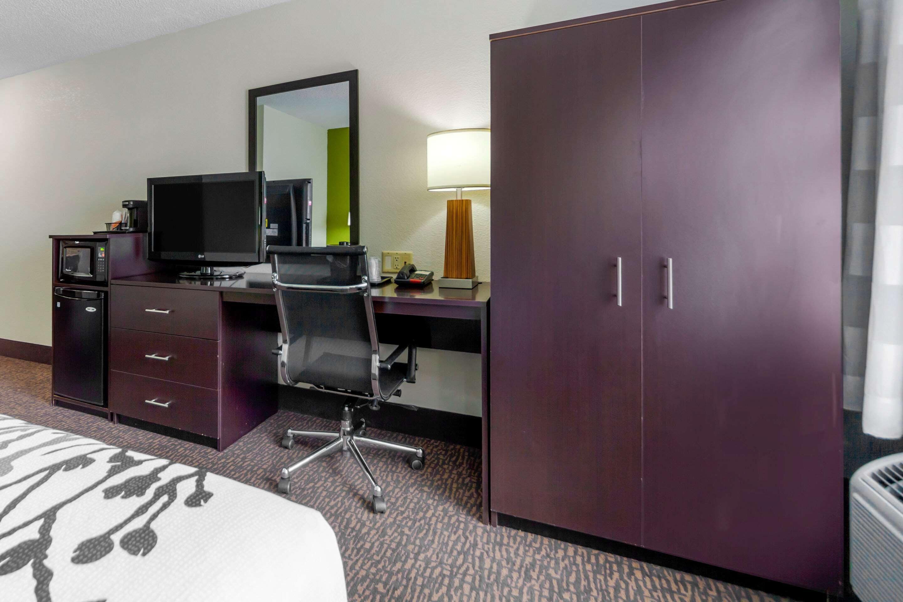 A hotel room with a bed, a desk with a computer, a chair, a wardrobe, a mirror, a lamp, and a microwave.