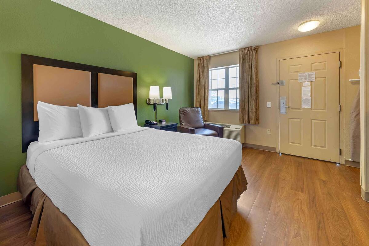 Habitación de hotel con una cama grande, un escritorio con silla, una lámpara y una ventana con cortinas.
