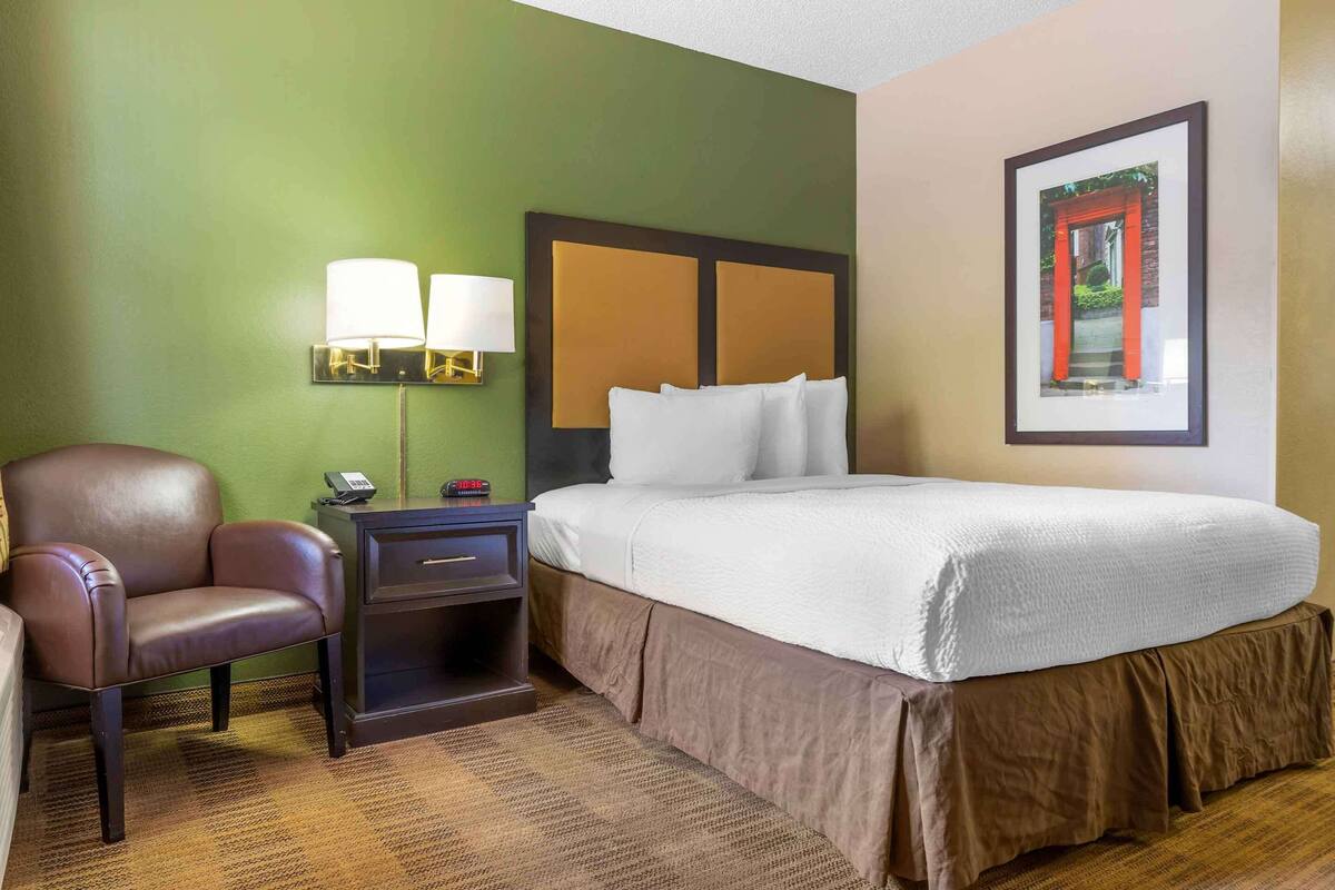 Una habitación de hotel con una cama, una silla, una mesita de noche y una lámpara.
