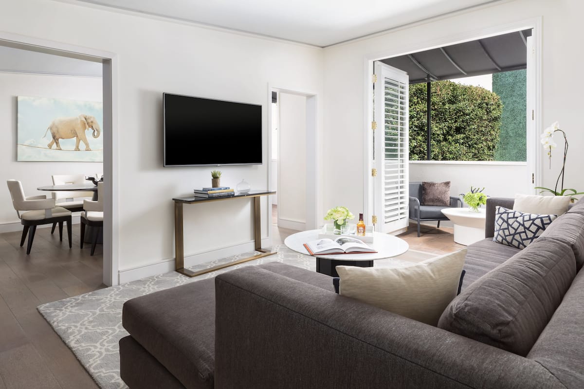 A modern living room with a grey sofa, a flat-screen TV, a dining area with a table and chairs, and a large window with a view of greenery.