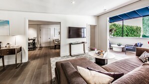 Un salon moderne avec un canapé, une table basse et un téléviseur. Il y a également un coin repas avec des chaises et une table. La pièce est dotée d’une grande fenêtre donnant sur un paysage verdoyant.