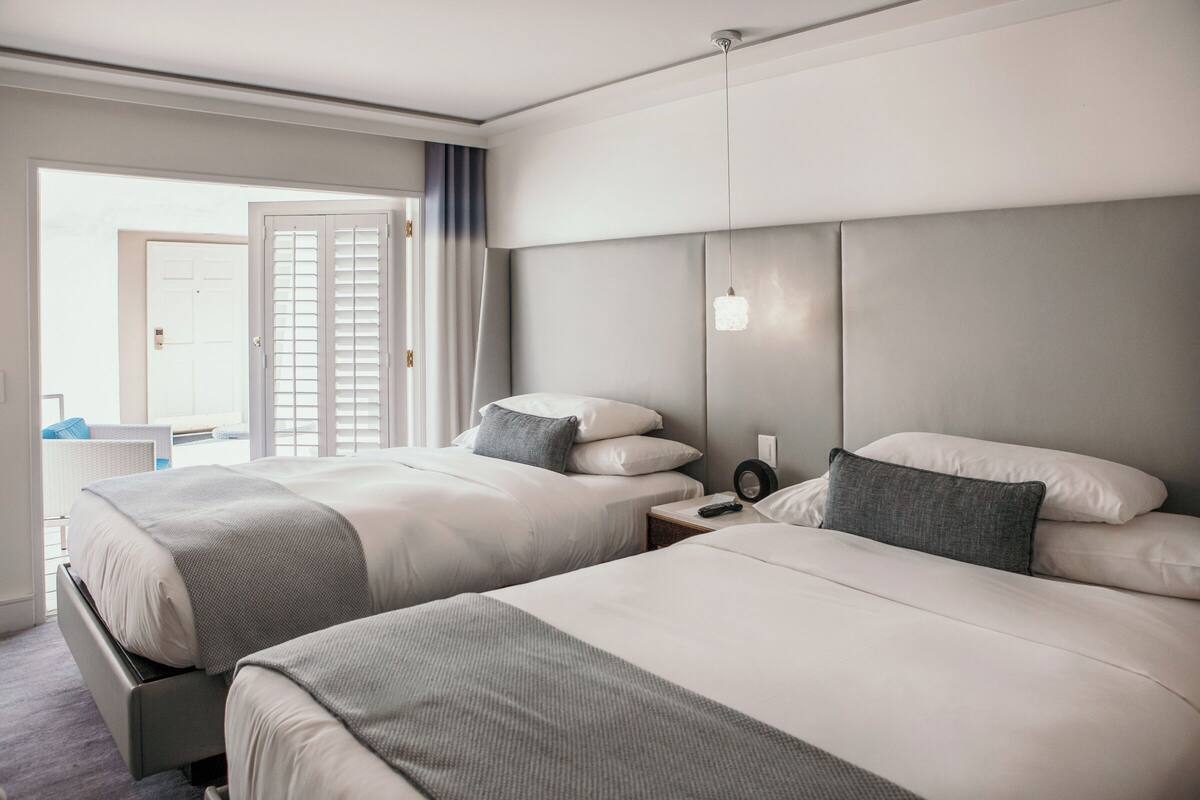 A hotel room with two beds, a nightstand, a lamp, and a view of the outside.