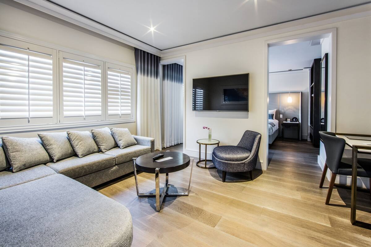 A modern living room with a sectional sofa, a round coffee table, and a grey armchair. A flat-screen TV is mounted on the wall, and there is a small table with a vase.