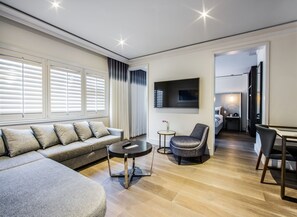 A modern living room with a sectional sofa, a round coffee table, and a grey armchair. A flat-screen TV is mounted on the wall, and there is a small table with a vase.