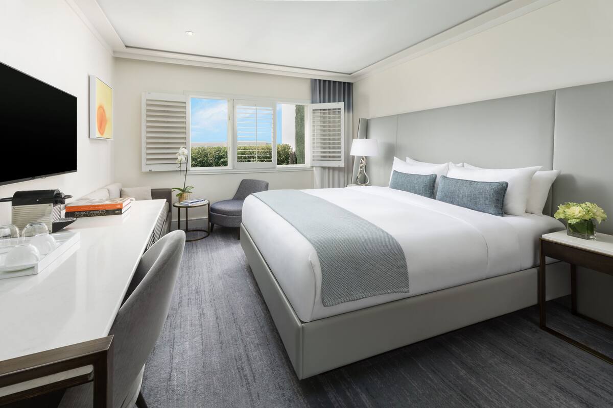A modern hotel room with a large bed, a desk with a flat-screen TV, a small round table, and a window with blinds.