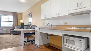 A modern kitchen with white cabinets, a microwave, and a sink. There is a desk with a chair and a lamp.