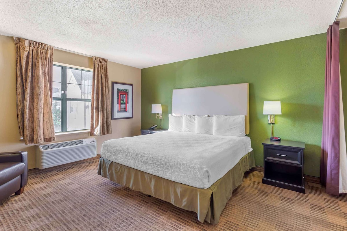 A hotel room with a large bed, two bedside tables with lamps, a chair, a window with curtains, and a framed picture on the wall.