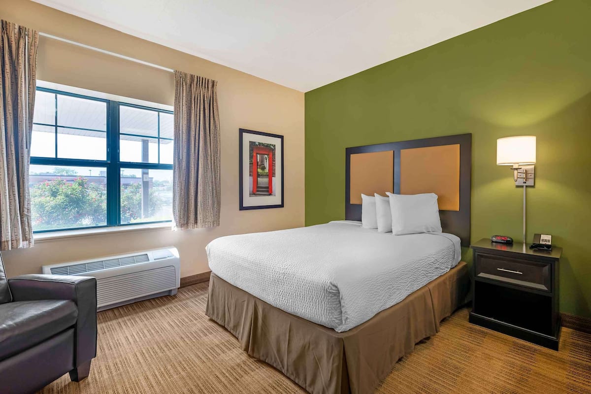 A hotel room with a large bed, a chair, a nightstand, a lamp, and a window with curtains.