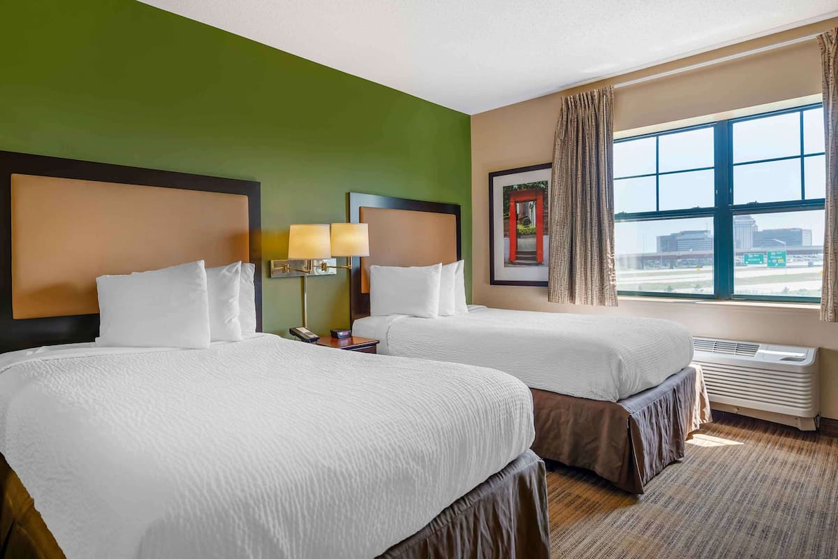 A hotel room with two beds, a large window with a view of the city, a wall-mounted lamp, and a framed picture.