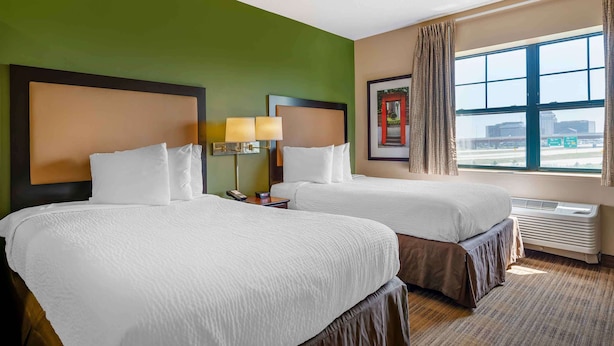 A hotel room with two beds, a large window with a view of the city, a wall-mounted lamp, and a framed picture.