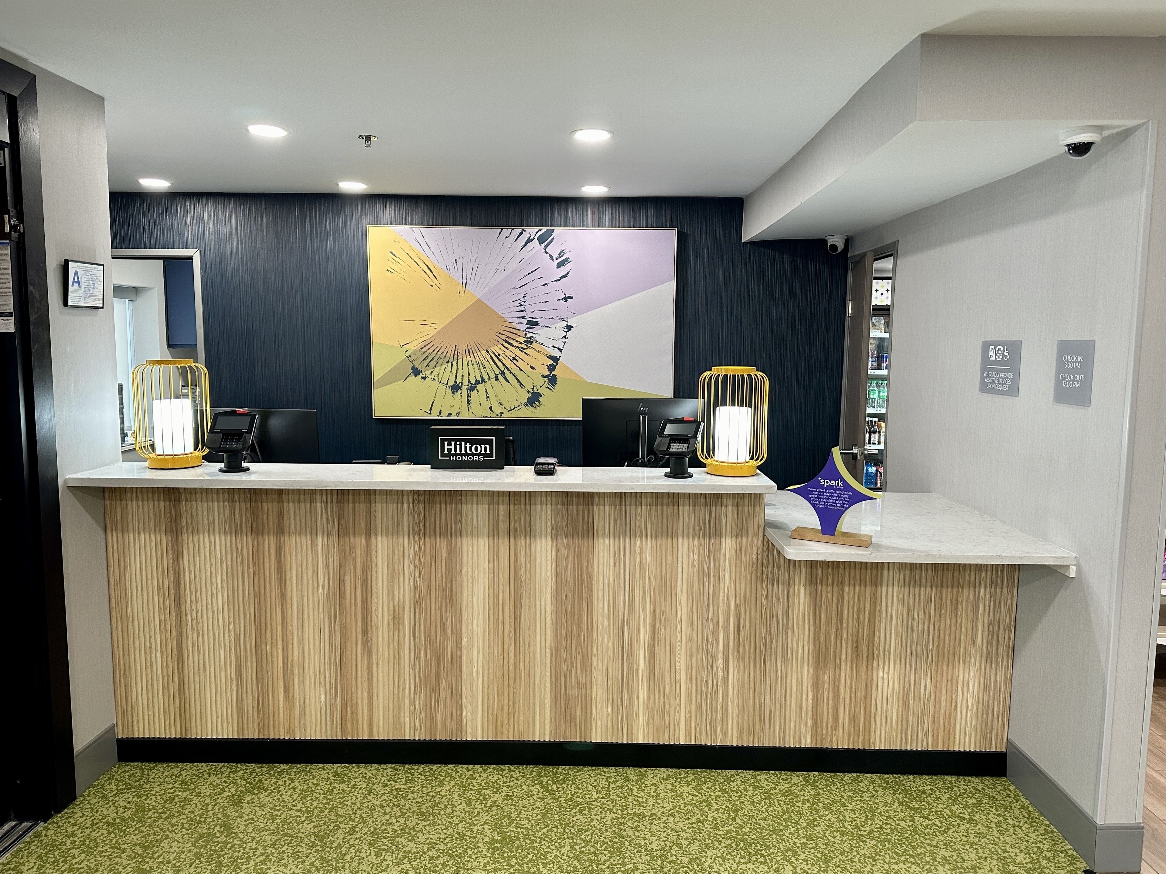 Un comptoir d’accueil en bois, une grande peinture abstraite au-dessus, deux lampes jaunes éclairées, un panneau « Hilton Honors », un panneau « spark » et une caméra de surveillance fixée au mur.